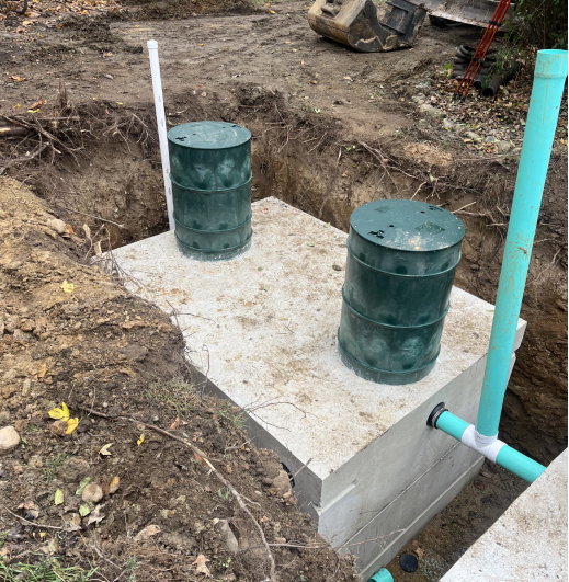 A concrete structure, possibly a septic tank or cistern, is partially buried in a dug-out area, featuring two green access lids, a vertical white vent pipe