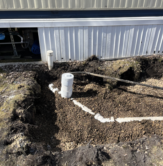 A dug-out area near a building featuring a white cylindrical pipe and gravel, indicating plumbing work in progress