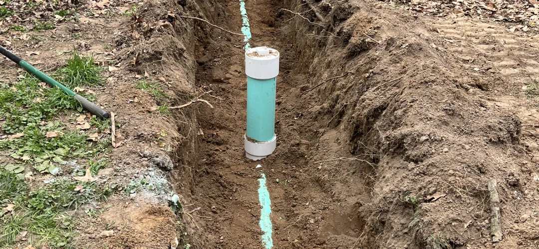 A long, narrow trench dug in a residential yard between a chain-link fence and a sidewalk, showing a teal pipe running along the bottom and a short