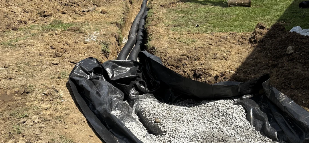 A trench dug in the dirt, lined with black plastic fabric and filled with small gray drainage gravel