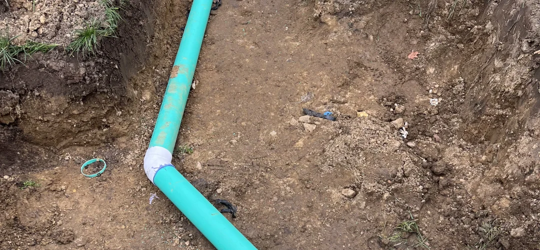 A trench dug in the dirt showing a bright teal pipe connecting with white PVC fittings, a white vent pipe (1)