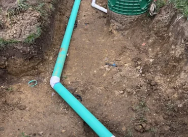 A trench dug in the dirt showing a bright teal pipe connecting with white PVC fittings, a white vent pipe (1)