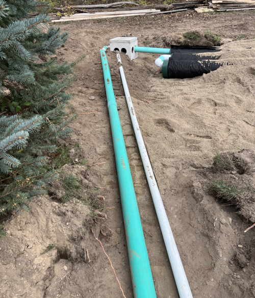 Two long pipes, one green and one gray, extend from a white drainage box into sandy ground, surrounded by greenery